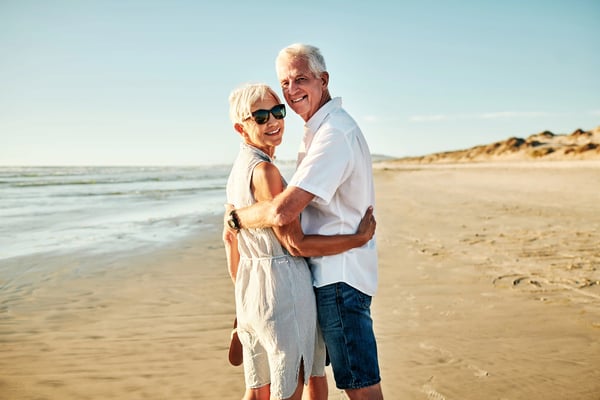 Älteres Paar umarmt sich lächelnd am Sandstrand mit Meer und Dünen im Hintergrund
