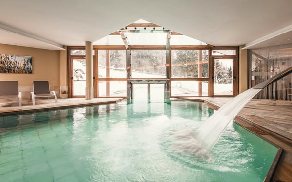 Innenpool mit Wasserfall, Liegen und grossen Fenstern mit Blick auf die winterliche Berglandschaft