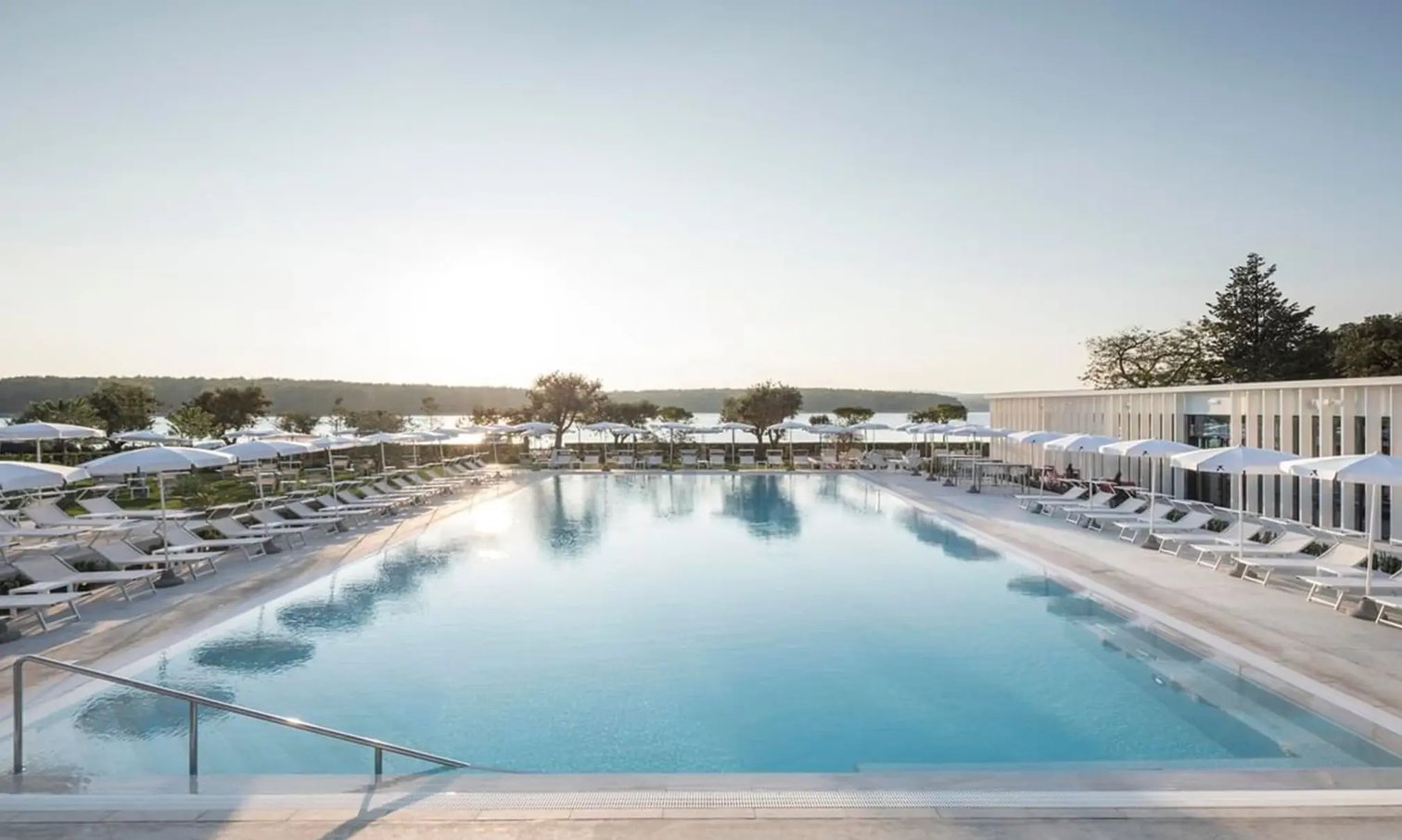 Grosszuegiger Aussenpool mit klarem Wasser, beidseitig angeordnete Sonnenliegen und Sonnenschirme, Blick auf Meer und flache Kuestenlandschaft bei ruhiger Stimmung