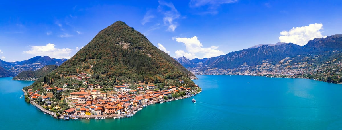 Luftaufnahme von Monte Isola mit Dorf am Ufer, steilem Hügel und blauem Iseosee