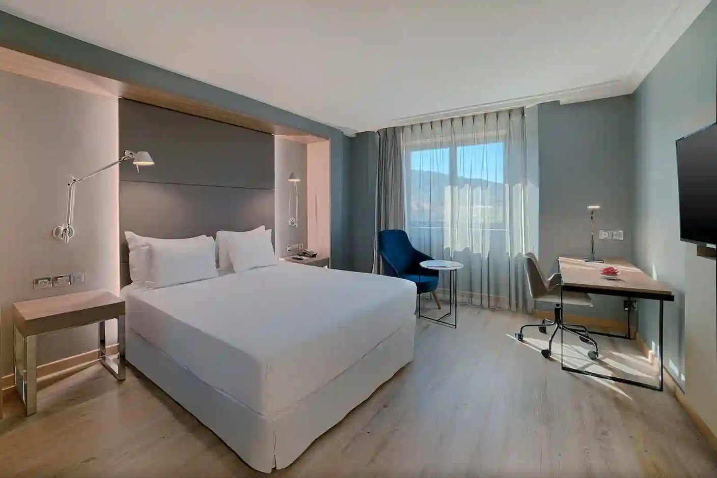 Modernes, helles Hotelzimmer mit Doppelbett, Schreibtisch, Sessel und bodentiefem Fenster, das viel Tageslicht in den Raum laesst.