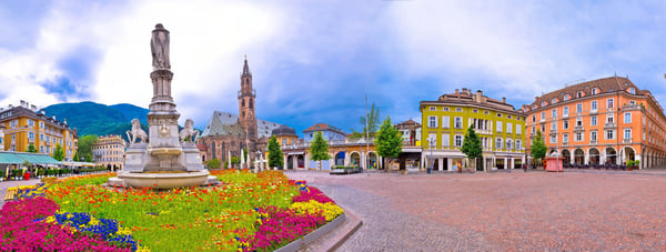 Panoramablick auf einen historischen Stadtplatz in Bozen mit bunten Gebäuden, Blumenbeeten, Brunnen und Bergkulisse im Hintergrund