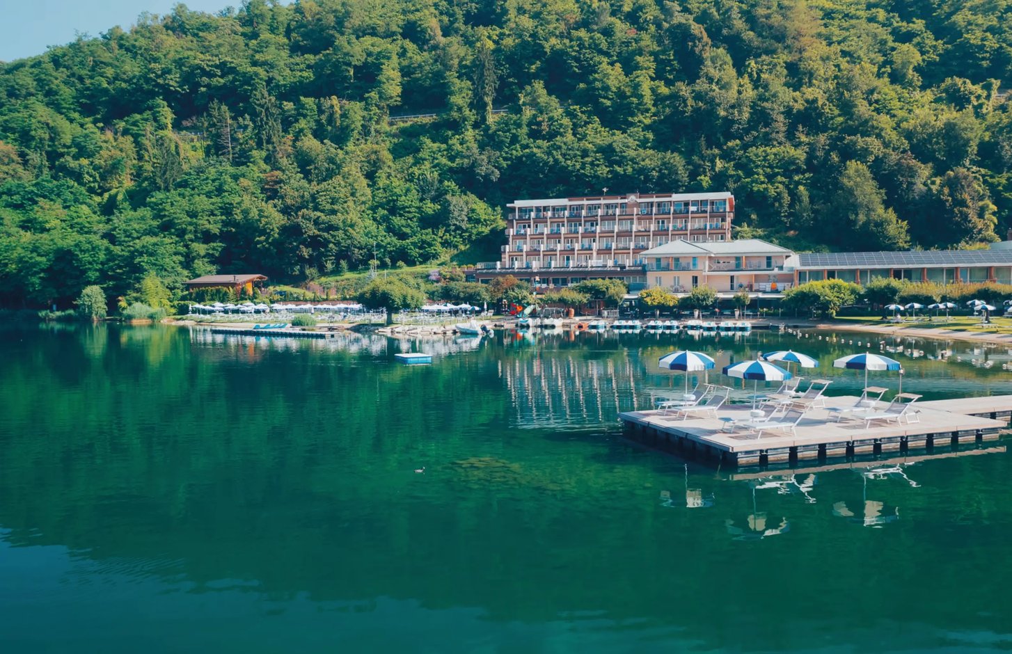 Hotelanlage am See mit Steg, Sonnenschirmen und Liegen auf schwimmender Plattform, umgeben von bewaldeten Hügeln.