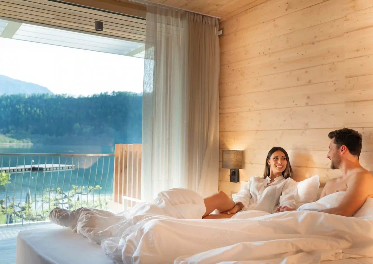 Paar liegt entspannt in einem modernen Hotelbett aus hellem Holz mit grosser Fensterfront und Blick auf einen See und bewaldete Berge