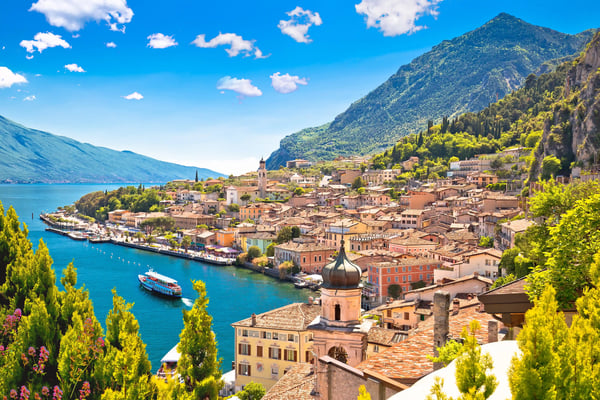 Blick auf den Ort Limone am Gardasee mit bunten Häusern, Kirchturm, Booten auf dem Wasser und steilen grünen Bergen unter blauem Himmel