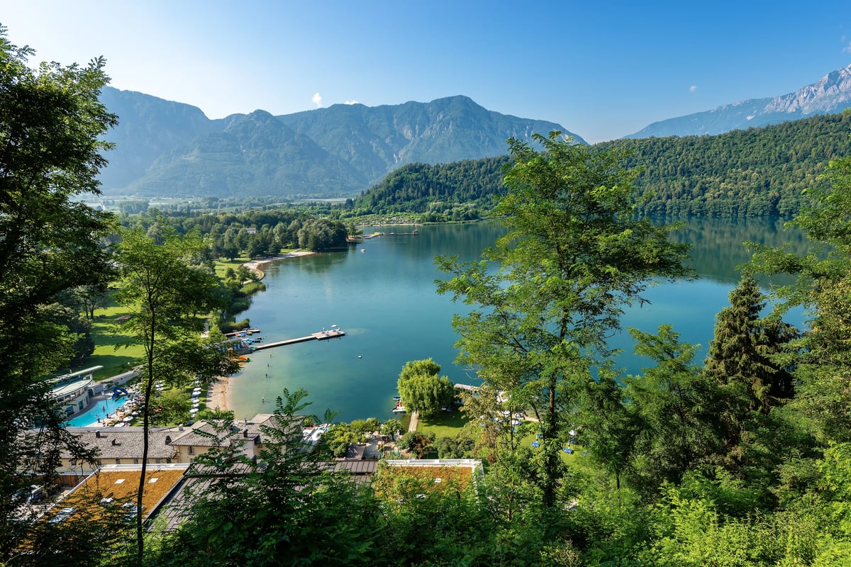 Panoramablick auf den Levico See mit grüner Uferlandschaft, Bergen und ruhigem Wasserulisse