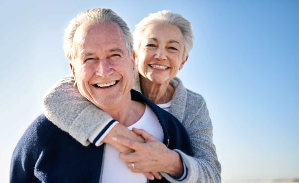 Älteres Paar lächelt in die Kamera, die Frau umarmt den Mann von hinten, beide stehen draussen vor blauem Himmel bei hellem Tageslicht