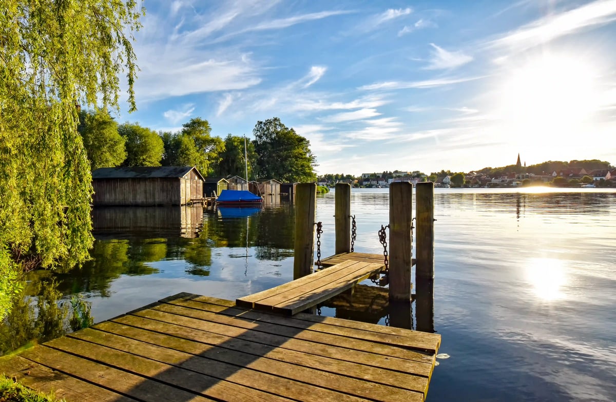 Holzsteg führt in einen ruhigen See, im Hintergrund kleine Bootshäuser, Bäume und Ortschaft im warmen Abendlicht