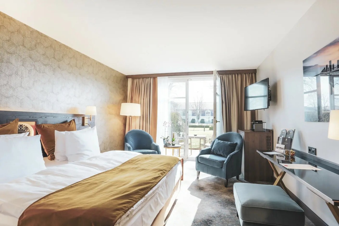 Helles Deluxe Hotelzimmer mit Doppelbett, gepolstertem Kopfteil, Sitzgruppe mit zwei Sesseln, Schreibtisch, Fernseher und bodentiefen Fenstern mit Blick in den Garten.