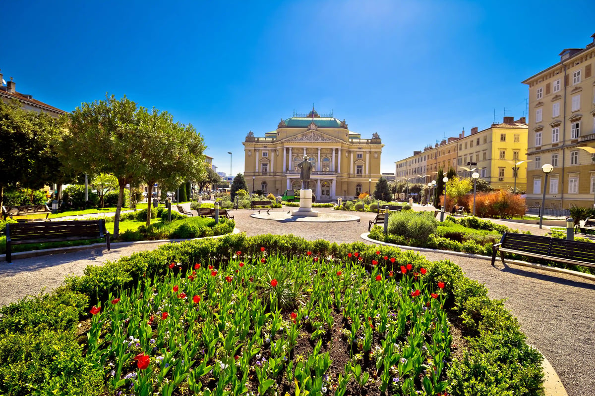 Nationaltheater in Rijeka mit gepflegtem Park, Blumenbeeten und klassizistischer Fassade bei blauem Himmel
