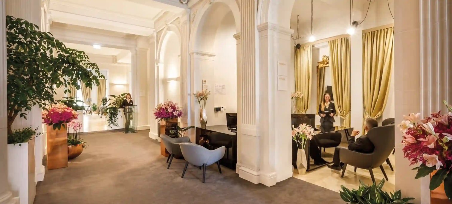 Elegante Hotellobby mit hohen Decken, weissen Saeulen, Rundboegen, stilvollen Sitzgruppen, Blumenarrangements und Personal im Gespraech mit Gaesten