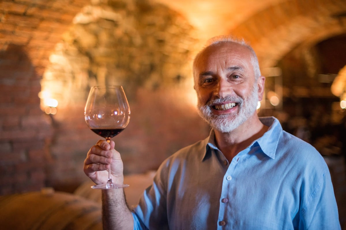 Winzer hält ein Glas Rotwein in einem Weinkeller im Piemont und lächelt in die Kamera