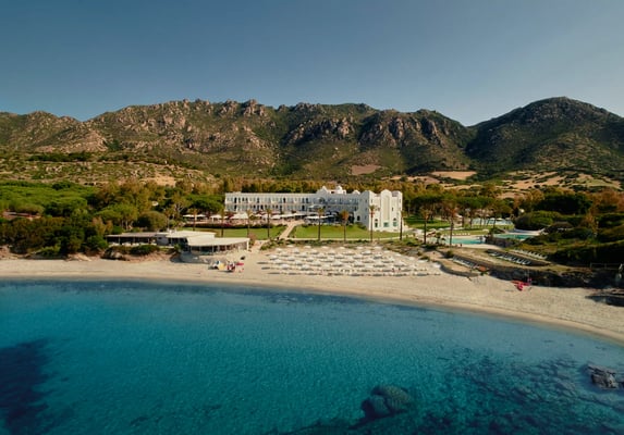 Luftaufnahme eines Hotels mit Privatstrand, Sonnenschirmen und türkisfarbenem Meer vor bergiger Landschaft auf Sardinien