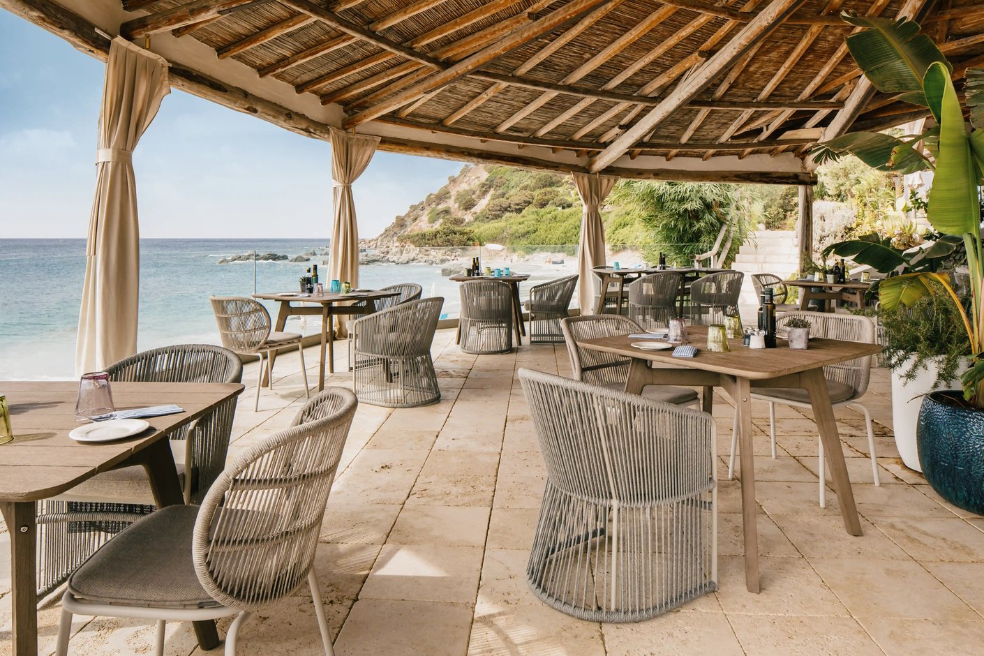 Offene Restaurantterrasse des Hotels Falkensteiner Capo Boi mit gedeckten Tischen, geflochtenen Stuehlen und Blick auf das ruhige Meer und die felsige Kuestenlinie
