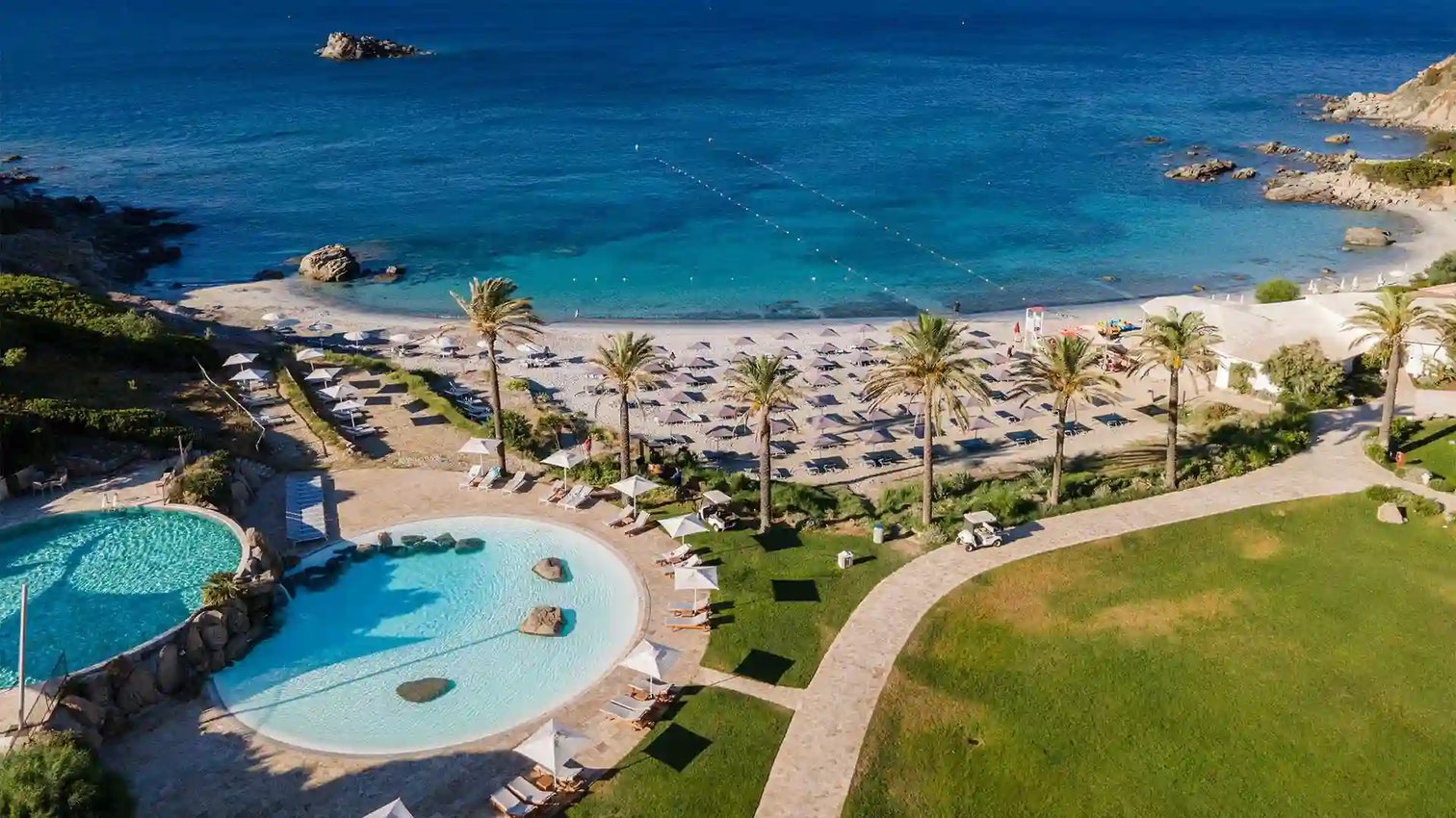 Luftaufnahme des Hotels Falkensteiner Capo Boi auf Sardinien mit weissem Gebaeudekomplex, direkter Strandlage, gepflegter Gartenanlage und Bergen im Hintergrund