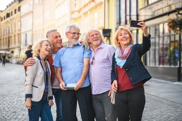 Fröhliche Gruppe älterer Reisender steht in einer Altstadtgasse und macht gemeinsam ein Selfie bei sonnigem Tageslicht