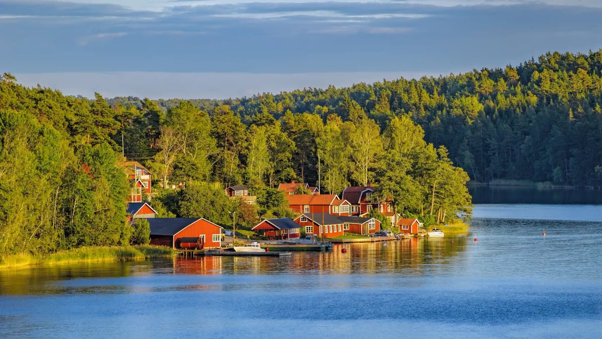 Rote Holzhäuser liegen idyllisch an einem ruhigen See, umgeben von dichtem gruenem Wald unter blauem Himmel