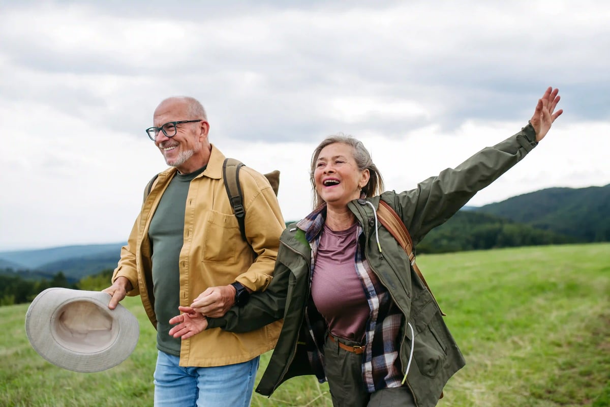 Lachendes Paar beim Wandern über eine grüne Wiese mit Rucksäcken und weiter Landschaft