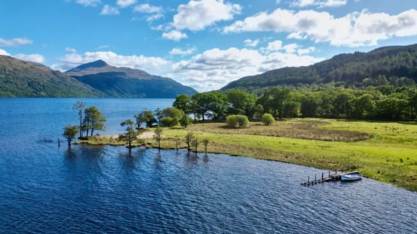 Loch Lomond mit ruhigem Wasser, bewaldetem Ufer und sanften Bergen unter blauem Himmel
