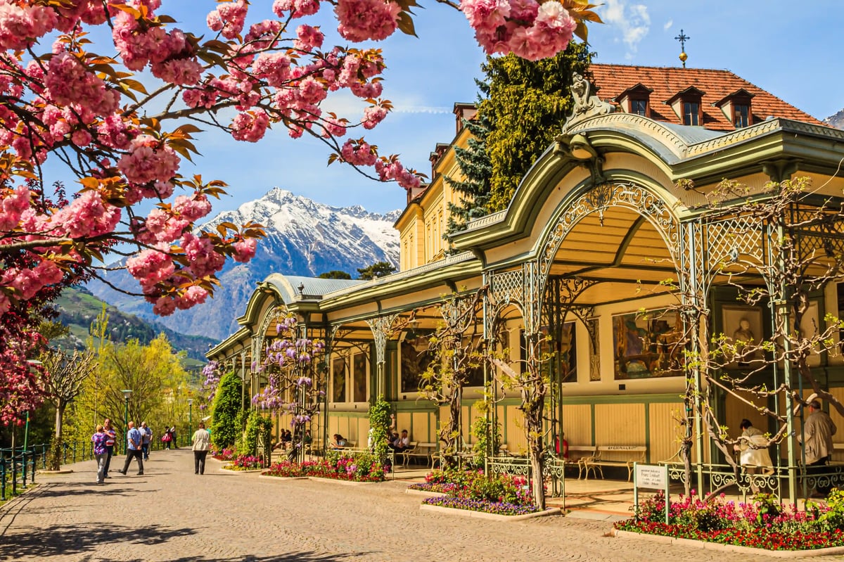 Wandelhalle in Meran mit Jugendstil Architektur, Blüten und schneebedeckten Bergen im Hintergrund