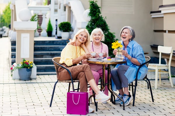 Drei ältere Frauen sitzen lachend an einem kleinen Tisch im Freien und trinken Kaffee