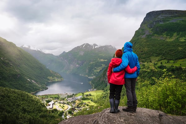 Paar steht auf einem Felsen und blickt auf den Geirangerfjord mit steilen Berghängen und tiefem Fjordwasser