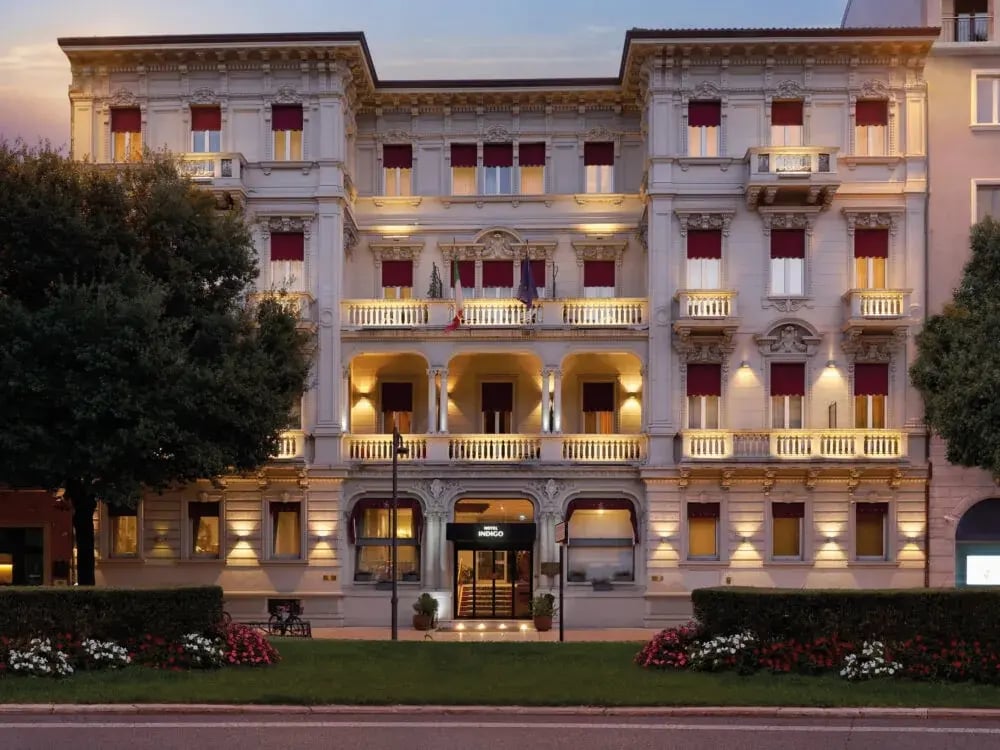 Beleuchtete Fassade des Hotel Indigo in Verona mit klassischer Architektur, Balkonen, roten Fensterlaeden und gepflegtem Vorgarten in der Abenddaemmerung.