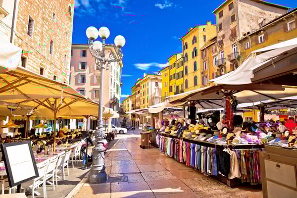 Lebendiger Marktplatz mit bunten Häuserfassaden, Marktständen, Cafes und Sonnenschirmen unter blauem Himmel