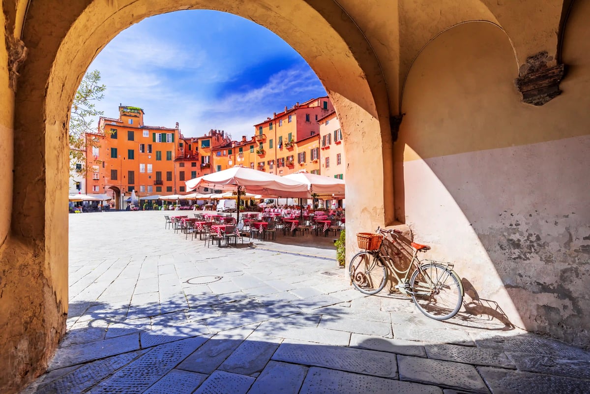 Vue à travers une arche en pierre sur une place ensoleillée avec des tables de café sous des parasols blancs, un vélo appuyé et des façades orange.