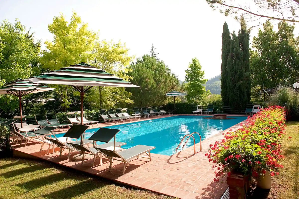Piscine extérieure rectangulaire à l’eau turquoise, entourée de chaises longues et de parasols rayés vert et blanc, avec des fleurs rouges et de grands cyprès.