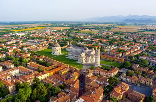 Luftaufnahme der Piazza dei Miracoli in Pisa mit Schiefem Turm, Dom und Baptisterium, umgeben von roten Ziegeldächern und grünen Rasenflächen.