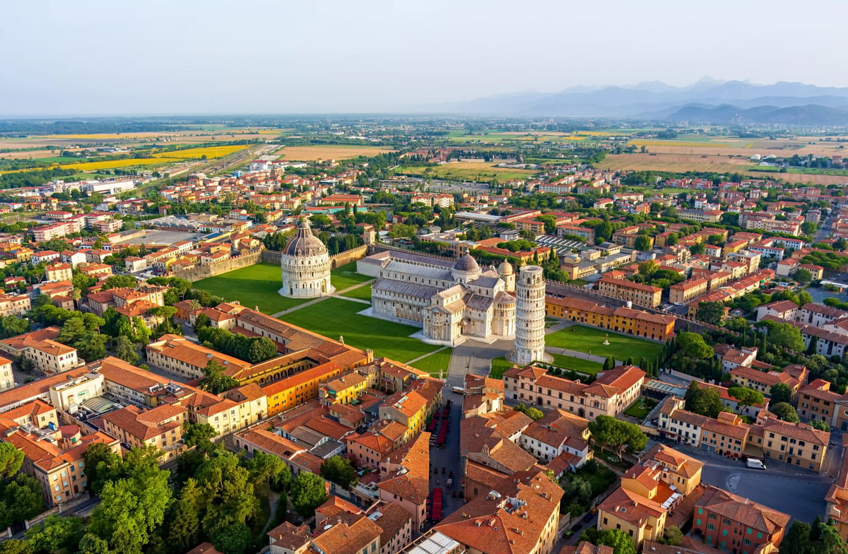 Vue aérienne de la Piazza dei Miracoli à Pise avec la tour penchée, la cathédrale et le baptistère, entourés de toits en tuiles et de pelouses.