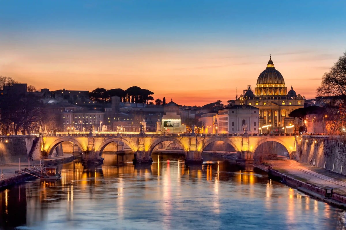 Blick auf den Tiber bei Nacht mit beleuchteter Brücke und Petersdom im Hintergrund unter farbigem Abendhimmel