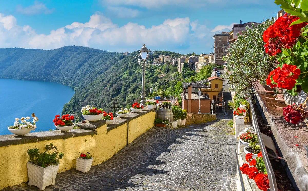 Malerischer Weg mit Blumen entlang einer Mauer mit Blick auf den Bolsenasee und bewaldete Hügel der Albaner Berge