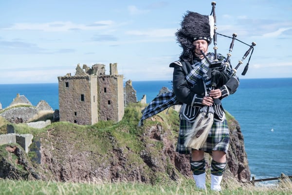 Dudelsackspieler in traditionellem Kilt steht auf einer grünen Klippe, im Hintergrund eine Burgruine und das blaue Meer