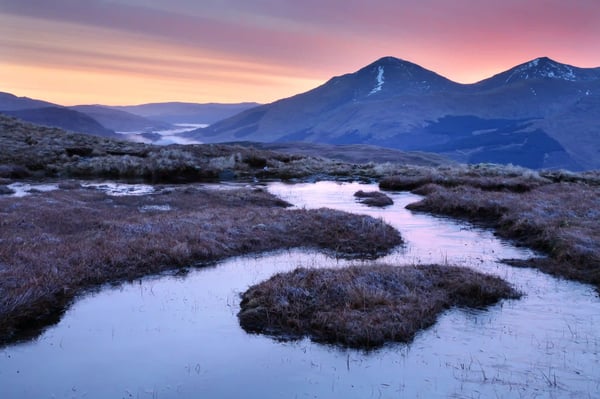 Moorlandschaft in den Trossachs mit gewundenem Bach im Vordergrund, sanften Hügeln und Bergen im Hintergrund bei farbigem Sonnenaufgang