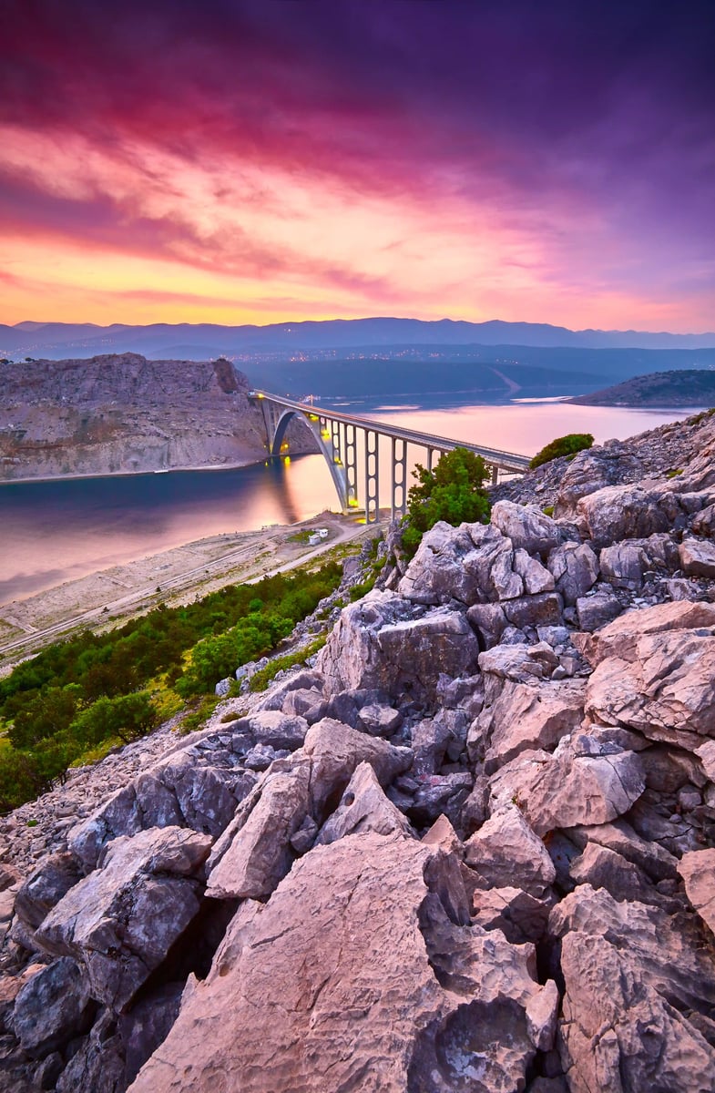Die Krk-Brücke überspannt bei Sonnenuntergang eine Meeresbucht, umgeben von felsiger Landschaft