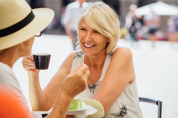 Zwei Frauen sitzen in einem Strassencafé, lachen miteinander und trinken Kaffee an einem sonnigen Platz.