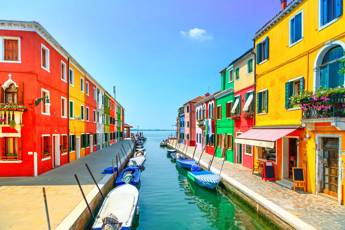 Vorderansicht eines Kanals auf Burano, Venedig, gesäumt von farbenfrohen Häusern (rot, gelb, grün, blau) und am Ufer vertäuten Booten.