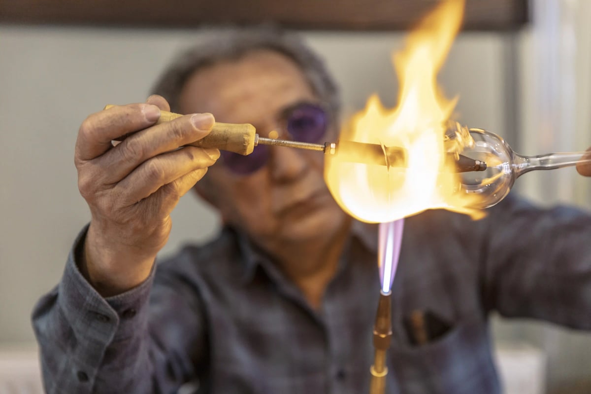 Nahaufnahme eines Glasbläsers, der ein Stück Glasrohr über einer großen, hellen Fackelflamme in seiner Werkstatt erhitzt.