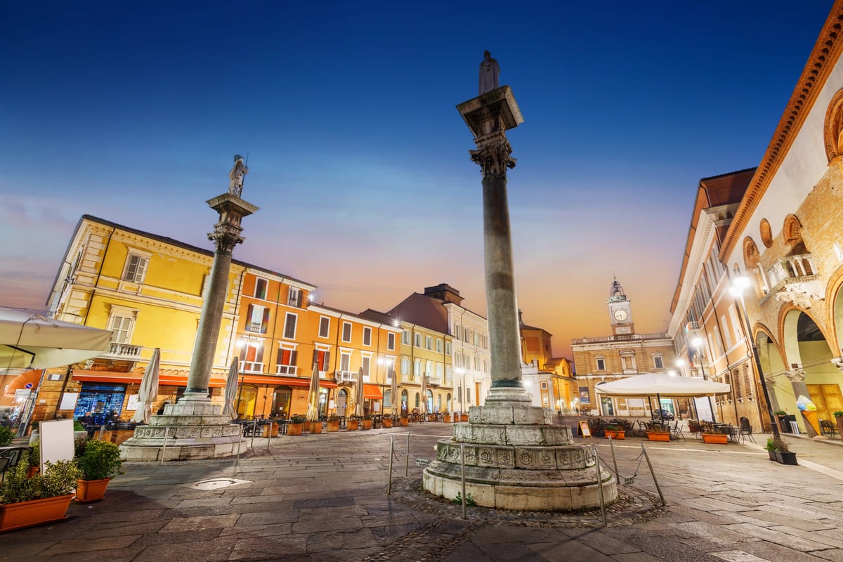 Blick auf einen historischen, gepflasterten Platz in Ravenna bei Dämmerung, mit zwei großen Steinsäulen, die Statuen tragen, und beleuchteten Cafés und Gebäuden unter einem dunklen Himmel.
