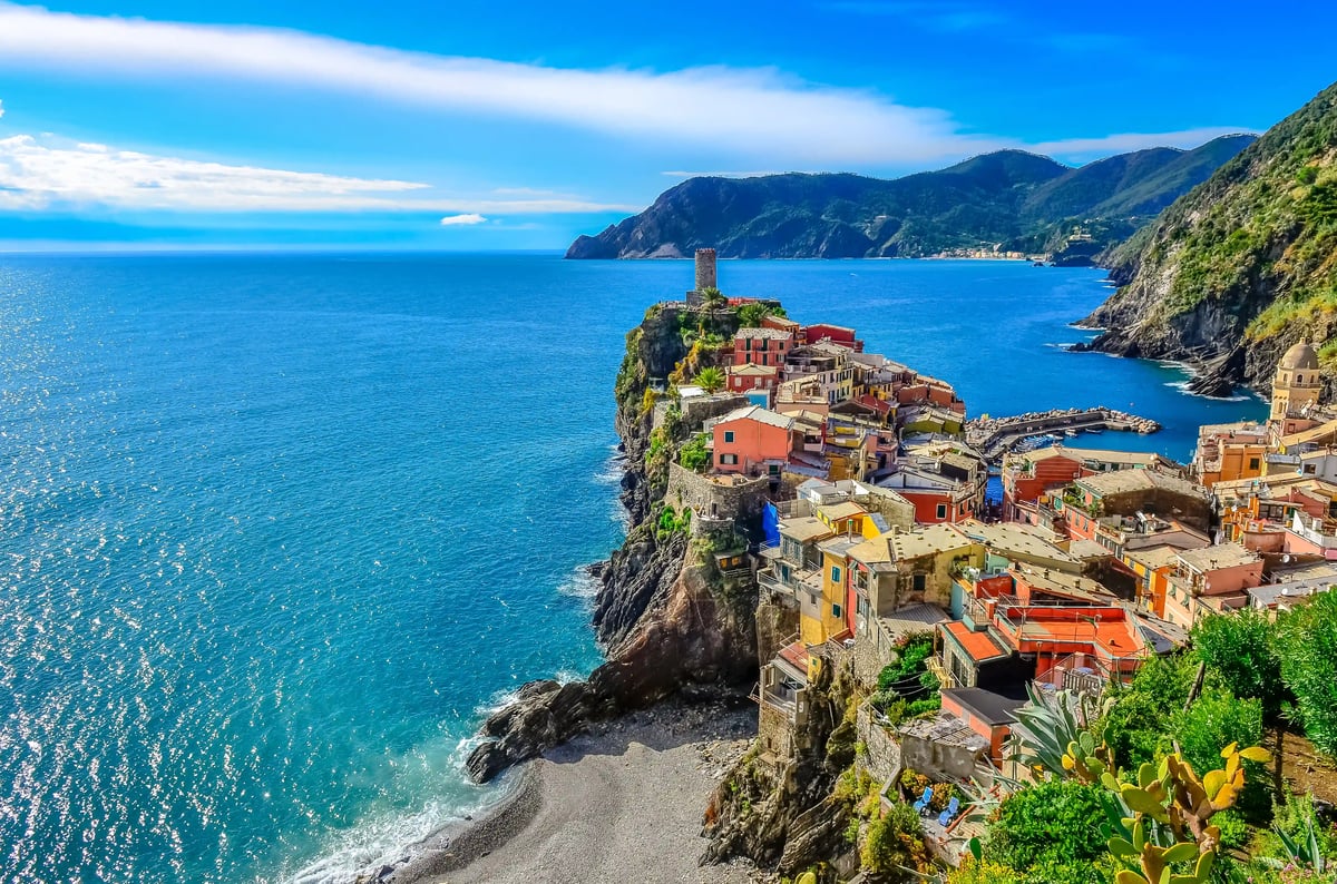 Maisons colorées et port de la Ligurie avec tour historique et drapeau italien, bateaux sur une mer calme devant des collines verdoyantes