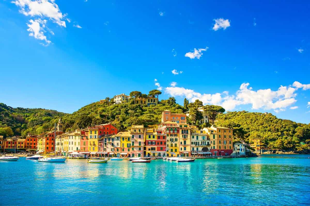 Maisons colorées et port de la Ligurie avec tour historique et drapeau italien, bateaux sur une mer calme devant des collines verdoyantes