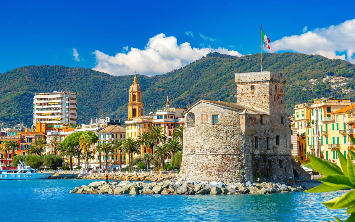 Château historique au port de Rapallo avec drapeau italien, promenade bordée de palmiers, bâtiments colorés et montagnes en arrière-plan