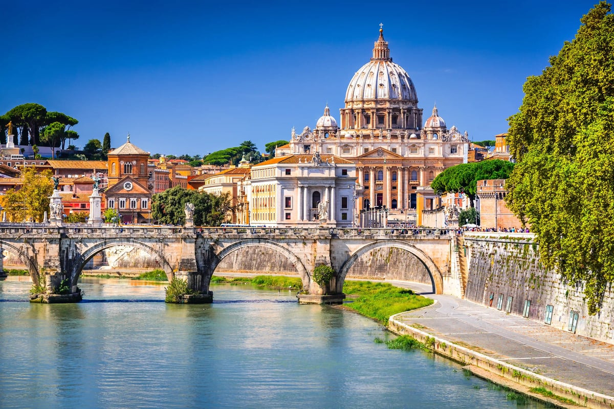 Petersdom in Rom erhebt sich hinter einer Steinbrücke über den Tiber bei klarem blauem Himmel