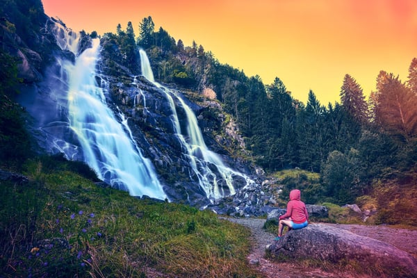 Grosser Wasserfall stürzt über dunkle Felsen in einem bewaldeten Tal, eine Person in roter Jacke sitzt auf einem Stein und blickt auf das fliessende Wasser