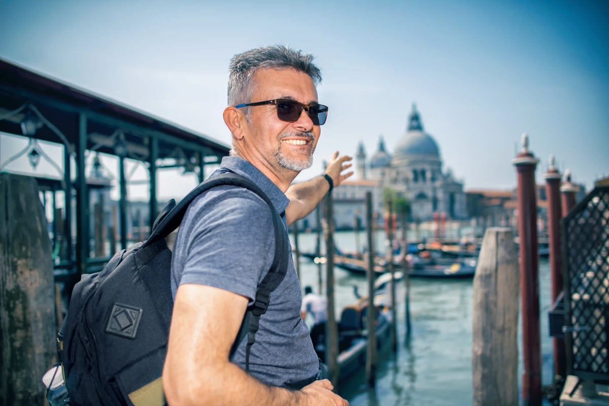 Lächelnder Mann mit Rucksack zeigt auf die Lagune von Venedig mit Booten und Kirche im Hintergrund