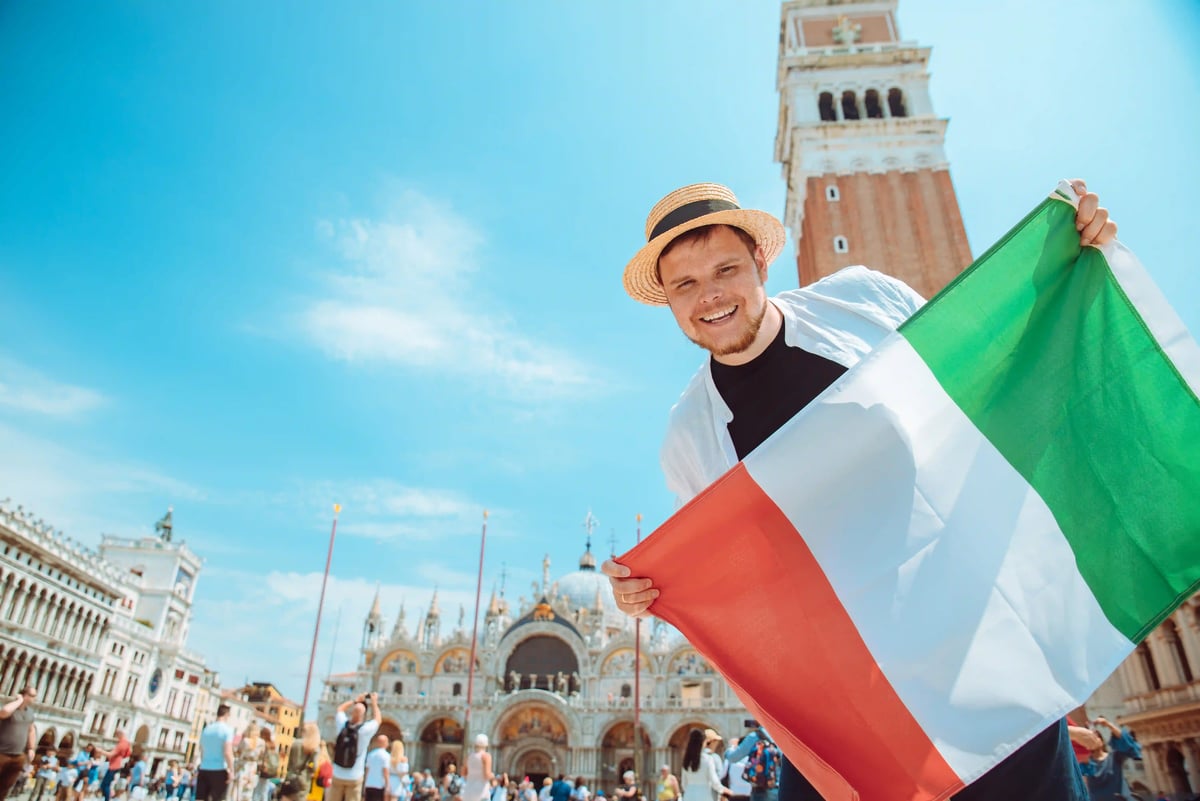 Lächelnder Mann hält eine italienische Flagge auf dem Markusplatz in Venedig mit historischem Gebäude im Hintergrund