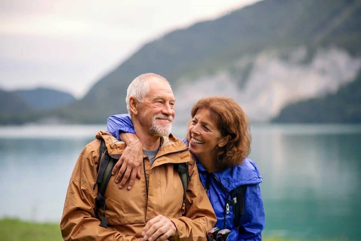 Älteres Paar in Outdoor Kleidung steht umarmt vor einem Bergsee und lächelt sich an