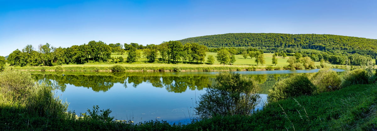 Paysage fluvial paisible de la rivière Doubs avec eau réfléchissante, prairies vertes et collines boisées sous un ciel bleu